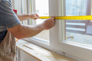 Un artisan qualifié utilisant un mètre ruban pour mesurer soigneusement les dimensions d'un cadre de fenêtre, debout devant un mur dans un atelier lumineux.